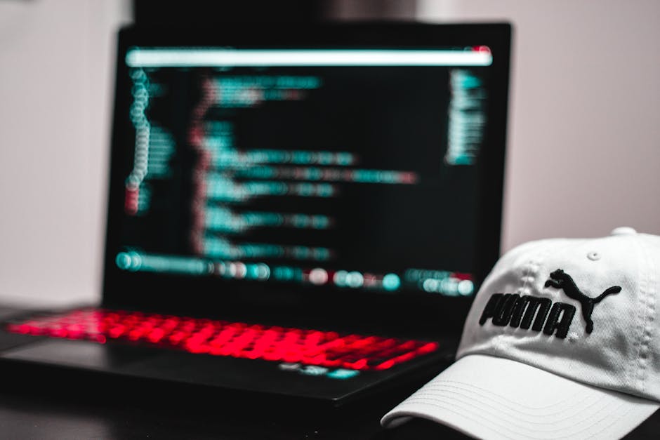 This image shows a slightly blurred laptop on a desk, displaying lines of code or text on its screen, with a backlit keyboard emitting red light in a dark environment. In the foreground, a white Puma-