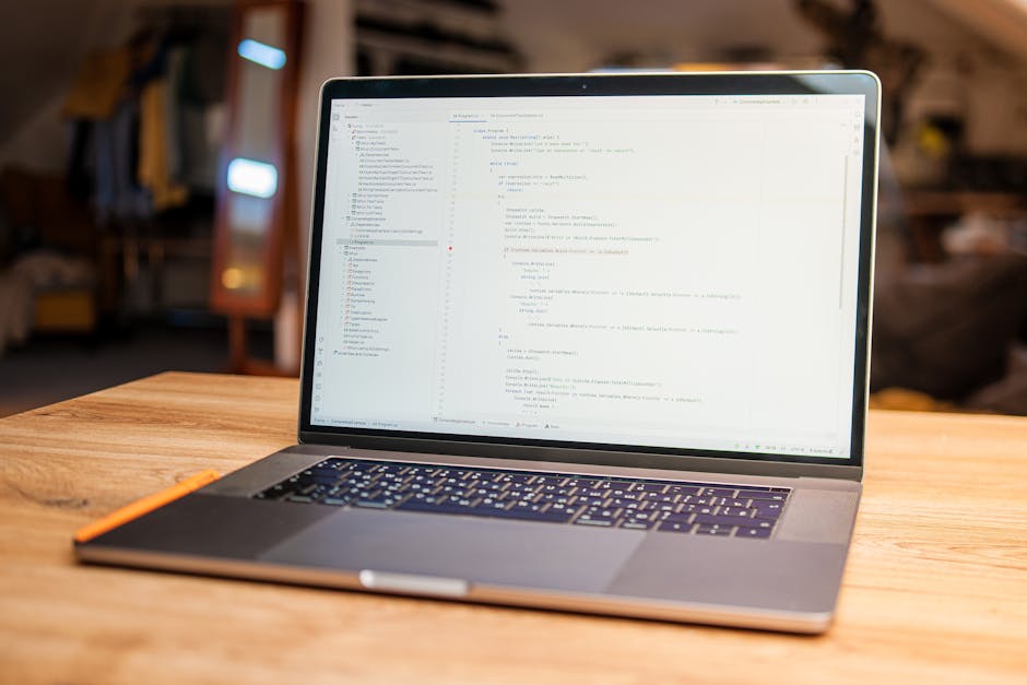 A silver laptop with a black keyboard sits open on a wooden desk, displaying lines of code or programming text on its screen. The background features a blurred indoor setting with warm lighting, woode